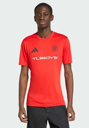 Ung mand iført en rød sports T-shirt med korte ærmer med teksten "TÜRKİYE", Adidas-logo og rundt emblem på brystet.