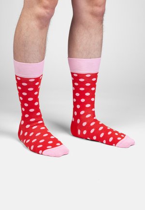 Ben iført røde sokker med lyserøde polkaprikker, lyserøde manchetter og lyserøde tæer, stående på en ensartet lysegrå overflade og baggrund.