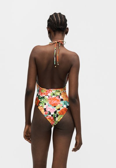 Kvinne med flettehår som har på seg en ryggløs, blomstrete heldrakt med oransje, rosa og grønne blomster på en rutete bakgrunn.