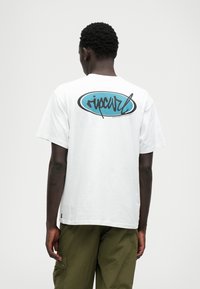 Biely bavlnený tričko s okrúhlym tyrkysovým logom na chrbte, obsahujúcim čierny text a písaný dizajn. Krátke rukávy, voľný strih.