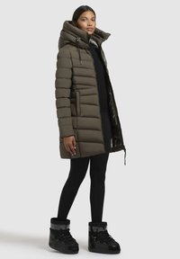 Olive green puffer coat with a large hood, quilted pattern, and front zipper. Paired with black leggings and high-top black boots.