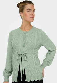 Svijetlozelena pletena cardigan jakna s kabelskim uzorcima, okruglim izrezom, pet dugmadi i remenom u struku. Dugi rukavi i detalj s valovitim rubom.