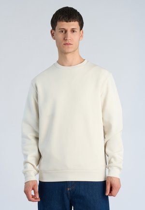 Junger Mann, der einen schlichten off-white Langarmpullover und blaue Jeans trägt, steht vor einem hellgrauen Hintergrund.