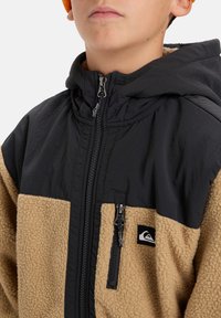 Giacca in fleece con parte superiore in nylon nero e parte inferiore in fleece beige, dotata di una zip frontale e piccolo logo ricamato sul lato sinistro.