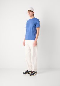 Blue cotton t-shirt with short sleeves, white loose-fit pants, and black and navy sneakers. Model wearing a light knit beanie.