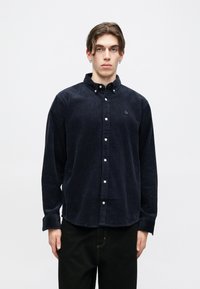 Ναυτικό πουκάμισο από χοντρό υφασμάτι με κλείσιμο ως τον γιακά, μακριά μανίκια, λευκά κουμπιά και μικρό κεντητό λογότυπο στο στήθος.