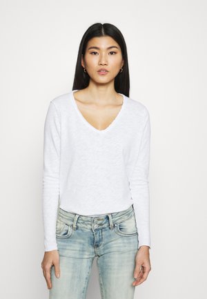 Hvid langærmet T-shirt med V-hals og rå kantdetaljer, parret med lyse denimjeans. Stoffet ser blødt og let struktureret ud.