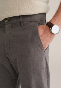 Bărbat îmbrăcat în pantaloni gri texturat, cu pulover bej din tricot, cu mâna în buzunar, și ceas argintiu cu brățară din piele maro.