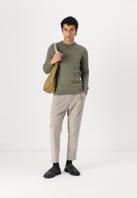 Zöld kötött pulóver, bézs pliszírozott nadrág, fekete cipő és világosbarna táska. A modellt fehér háttér előtt áll.