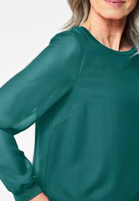Blusa verde a maniche lunghe con una texture liscia e scollatura arrotondata, dotata di una tasca discreta sul davanti e polsini aderenti.