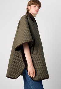 Gequilted oli-groen poncho met een bruine kraag, zijzakken en een bruine bies langs de randen. Textuurafwerking met een losse pasvorm.