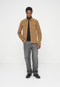 Veste-chemise en velours côtelé marron avec deux poches poitrine, associée à un col roulé noir et un jean gris à coupe droite, complétée par des bottes noires.