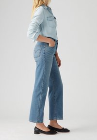 Camisa de denim azul claro con mangas enrolladas, combinada con jeans rectos azules que presentan costuras en los bolsillos traseros. Unos zapatos planos negros completan el look.