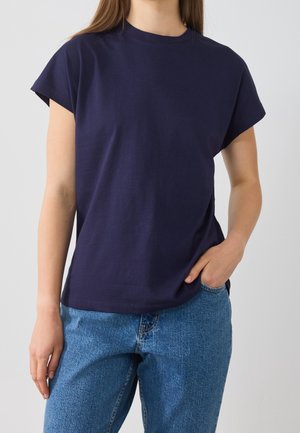 Kobieta ubrana w granatowy krótki rękaw t-shirt i niebieskie dżinsy, z jedną ręką w kieszeni na jednolitym tle.