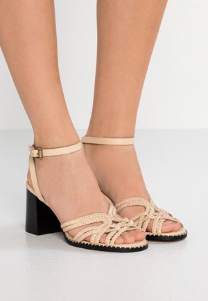 Pieds portant des sandales beiges à brides tressées, talons blocs et brides à la cheville, posés sur un sol lisse gris clair.