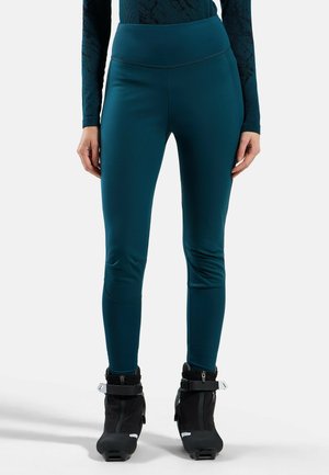 Woman wearing dark teal fitted leggings and black ski boots standing against a white background.
