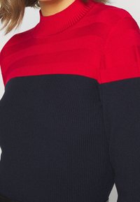 Maglione a maglia con un alto collo a coste. Presenta strisce orizzontali rosse e blu navy. Il materiale sembra morbido con una texture a coste.