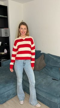 Γυναίκα που χαμογελά φορώντας πουλόβερ με κόκκινες και λευκές ρίγες, ανοιχτό μπλε τζιν και αθλητικά παπούτσια, όρθια δίπλα σε έναν μπλε καναπέ σε ένα σαλόνι.