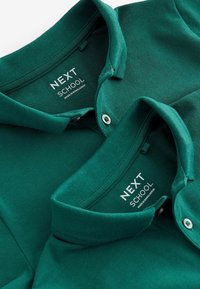 Camisas polo verdes apiladas, con tejido texturizado, detalle de cuello y botones. Las etiquetas dicen "NEXT SCHOOL", indicando el fabricante.