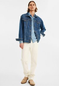 Tummansininen farkkutakki, jossa on napitettava sulku, kaksi rintataskua ja valkoinen aluspaidat, yhdistettynä kermaisiin housuihin ja beigeihin saappaisiin.