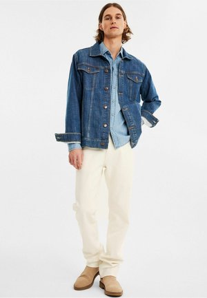 Dunkelblaue Jeansjacke mit Knopfverschluss, zwei Brusttaschen und weißem Unterhemd, kombiniert mit cremefarbener Hose und beigen Stiefeln.
