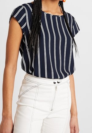 Vrouw draagt een marineblauwe blouse met witte verticale strepen en een witte broek met hoge taille, zichtbare stiksels en een rits aan de voorkant.