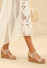 Wedge sandals with shimmering straps, cork wedge heel, and textured beige tones, paired with a sheer white lace cover-up and a woven bag.