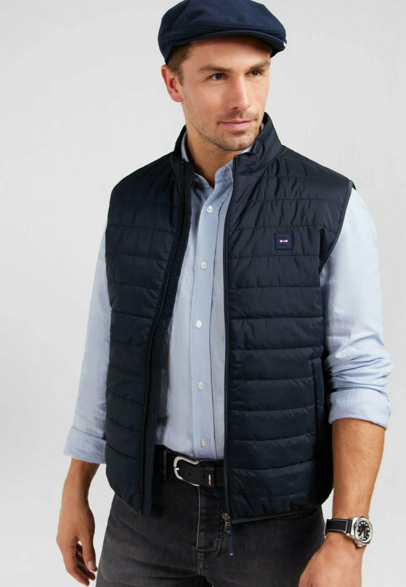 Gilet matelassé bleu marine avec fermeture éclair à l'avant, texture matelassée et patch logo. Porté par-dessus une chemise bleu clair à boutons et un jean foncé. Accessoirisé avec une ceinture et une montre.