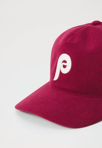 Burgunderfarget kordfløyelslue med en hvit brodert logo. Har en buet skjerm og teksturert overflate som fremhever stoffet og designet.