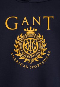 Haut à capuche bleu marine avec texte et logo jaunes. Présente un emblème circulaire avec une couronne, un design de laurier, et les mots "GANT AMERICAN SPORTSWEAR."
