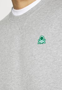 Grå sweatshirt gjord av bomullsblandning, med rund halsringning och en liten grön broderad bläckfisklogga på bröstet.