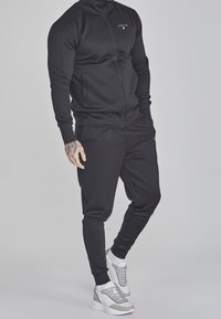 Chandal atlético negro hecho de tela suave, que presenta una chaqueta con cremallera y bolsillos laterales, y pantalones ajustados a juego con puños acanalados. Zapatillas plateadas.