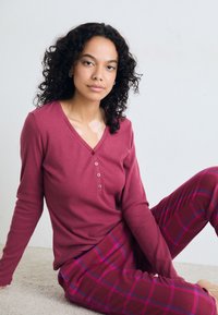 Långärmad, v-ringad topp i ribbat vinrött tyg med fyra knappar. Rutig pyjamasbyxor i vinröda, rosa och blå mönster.