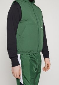 Man som bär en grön Nike Air ärmlös jacka över en svart långärmad tröja och gröna träningsbyxor med vita och svarta sidostreck.