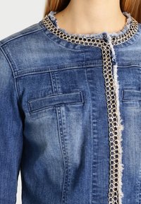 Giacca di jeans azzurro chiaro con colletto sfrangiato e dettagli in catena d'argento lungo la chiusura frontale, con due tasche sul petto.
