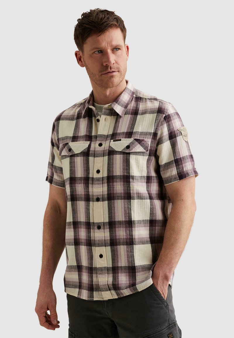 Homme portant une chemise à manches courtes à carreaux avec des poches poitrine, debout avec une main dans la poche devant un fond uni.