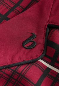 Red and black plaid fabric with a smooth texture, featuring a black embroidered number "6" and contrasting black piping along the edge.