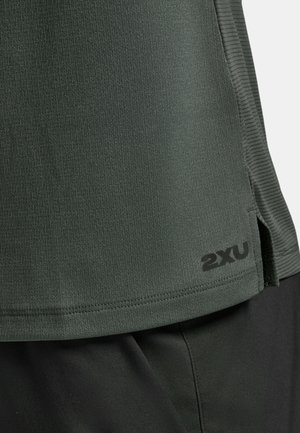 Close-up van de zoom van een donkergroen gestructureerd sportshirt met het "2XU"-logo boven een zwarte korte broek, met zichtbare zijnaden en een kleine stofsplitsing.