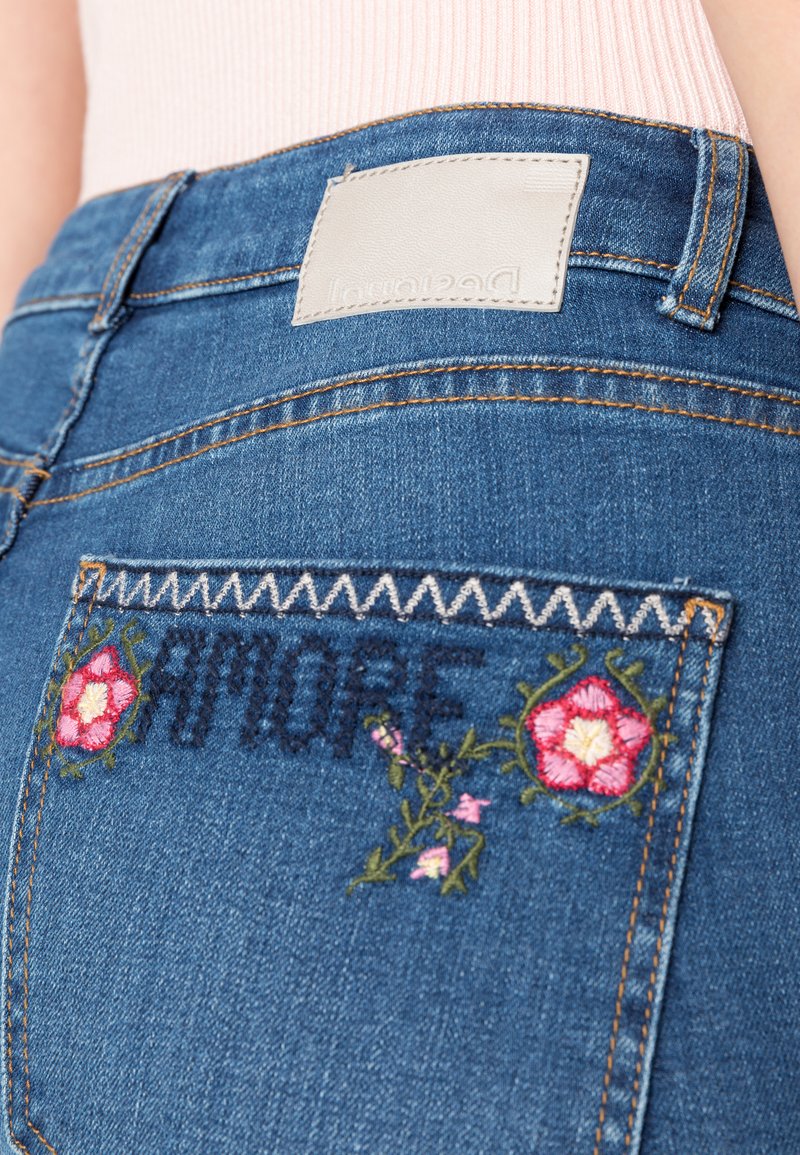 Die Jeans bestehen aus einem blauen Waschungsstoff und verfügen über eine Gesäßtasche, die mit "AMORE" in dunklem Faden bestickt ist, akzentuiert von floralen Designs in Pink und Grün.