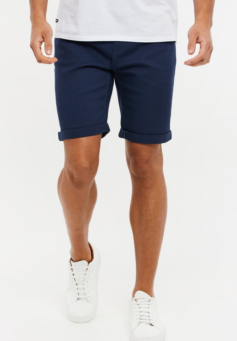 Homme portant un short roulé bleu marine, des baskets blanches et un t-shirt blanc, se tenant devant un fond blanc uni.