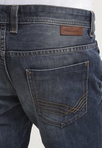 Jeans de mezclilla azul oscuro con un corte regular. Presentan una etiqueta de cuero, sutiles detalles de costura en los bolsillos y un diseño clásico de 5 bolsillos.