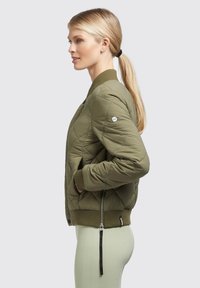 Olivovo zelená prešívaná bomber bunda s rebrovanými manžetami a lemom, so zipsom na bočnej strane a detailom bočného zipsu.