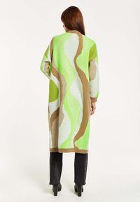 Long knit cardigan with wave patterns in green, brown, and white. Features drop shoulders and ribbed cuffs, worn over dark pants.