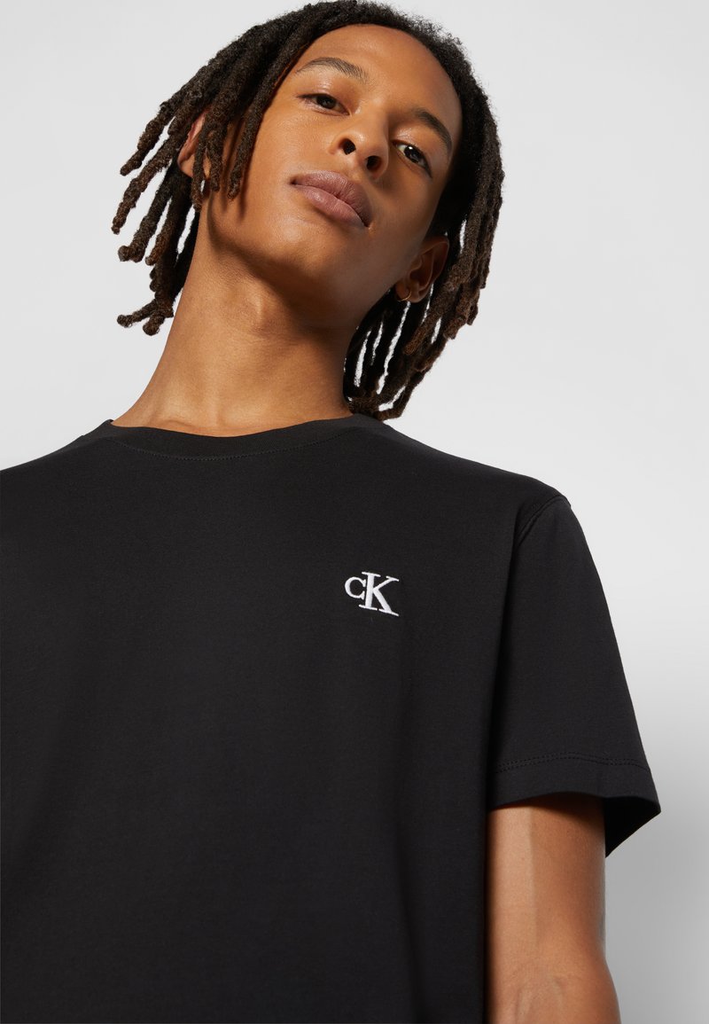 Persona joven con rastas hasta los hombros que lleva una camiseta negra con un pequeño logo blanco "cK" en el pecho sobre un fondo liso.