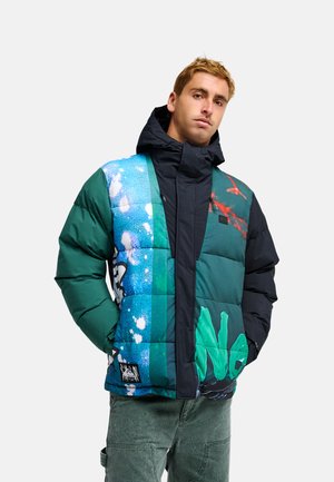 Homme portant une veste matelassée colorée avec des motifs abstraits bleus, verts et noirs et un pantalon cargo vert, debout les mains dans les poches.