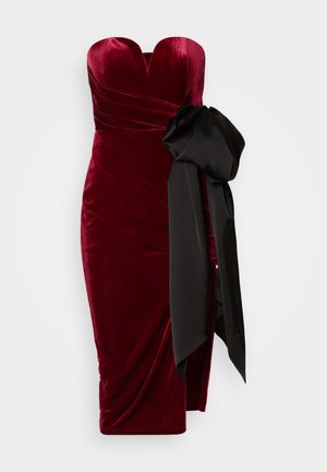 Burgundi, bársonyos ruha szív alakú dekoltázzsal, redőzött kialakítással és egy nagy fekete selyem masnival az oldalon. Midi hosszúságú, testéhez simuló szabással.