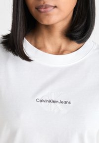 T-shirt bianca in cotone con collo rotondo e logo ricamato "CalvinKleinJeans" in grigio, con una texture sottile del tessuto.