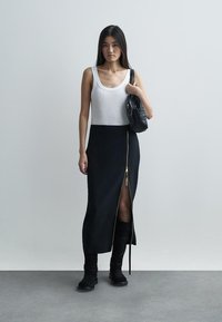 Czarna spódnica z zamkiem z przodu, w połączeniu z białym topem na ramiączkach. Modelka nosi czarne kozaki do kolan i trzyma małą czarną torebkę.