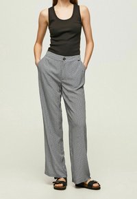 Frau in gemusterten, weit geschnittenen Hosen, schwarzem ärmellosem Oberteil und schwarzen Sandalen mit Korksohle, steht mit den Händen in den Taschen.