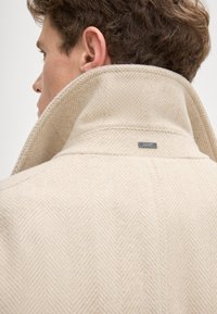 Beiger Fischgrät-Wollmantel mit hochstehendem Kragen, der ein Metall-Logo auf der Rückseite trägt und seine anspruchsvolle Textur und Gestaltung hervorhebt.